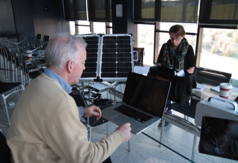 A portable solar panel and battery setup being demonstrated indoors with a laptop.
