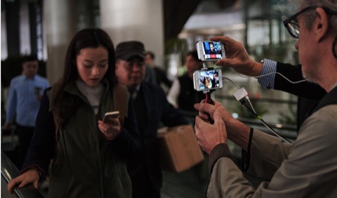 A camera operator filming on location with a dual-phone rig as a crowd passes by.